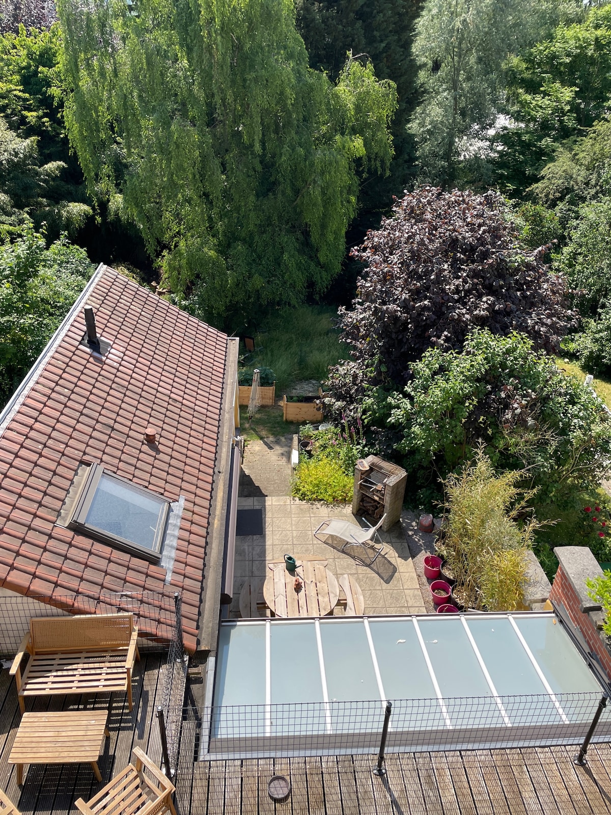 An aerial view showcases a terraced outdoor area featuring a patio with a glass covering. Surrounding greenery includes various trees and shrubs, creating a natural enclosure. A small structure for barbecuing is visible, along with a few seating options arranged on the patio.