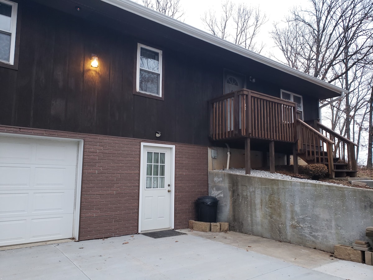 A two-story building is depicted, featuring a light-colored garage door and a front entryway with a wooden railing. The entrance is surrounded by trees, creating a natural setting, and steps lead up to the main door, enhancing accessibility.