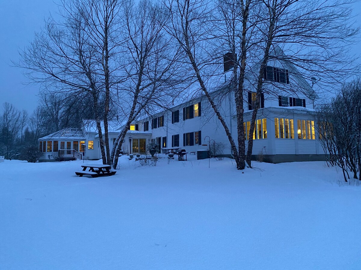The farmhouse inn is set against a snowy landscape, showcasing its multiple windows illuminated in the evening light. Leafless trees frame the building, and a picnic table can be seen in the foreground, all surrounded by a soft blanket of snow.