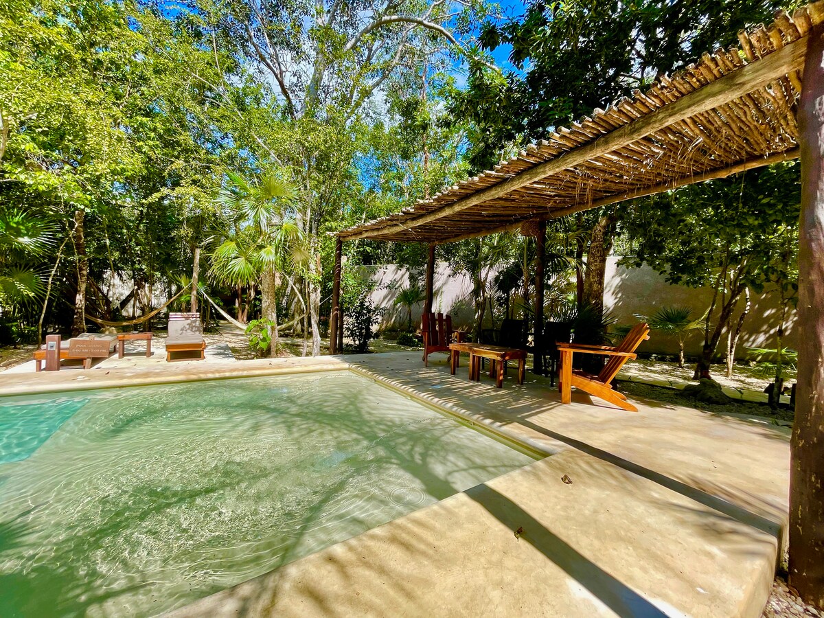 A shaded outdoor area is showcased, featuring a refreshing pool surrounded by lush greenery. Wooden lounge chairs and a dining table with several chairs provide comfortable spaces for relaxation and enjoyment in a tranquil jungle setting.