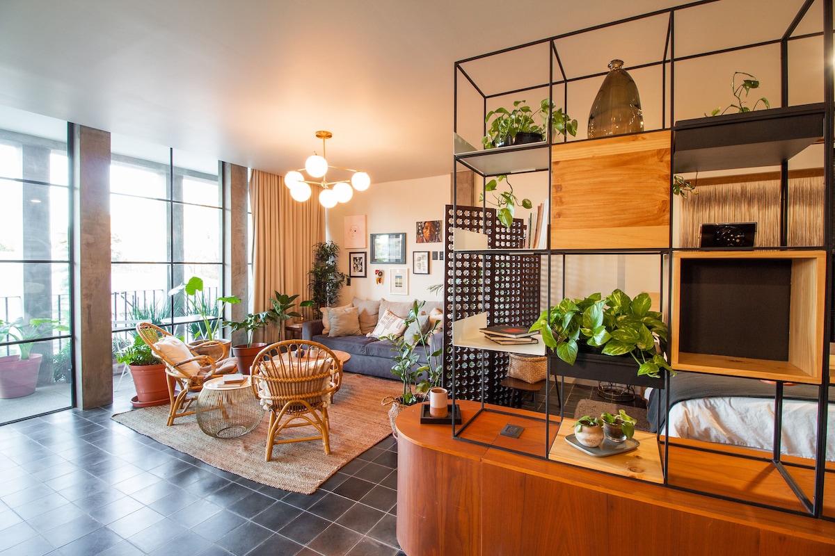 A spacious living area features large windows that invite natural light, complemented by indoor plants and contemporary furniture. A comfortable sofa is positioned next to a decorative room divider, showcasing an assortment of greenery. The open layout provides a welcoming atmosphere for relaxation.
