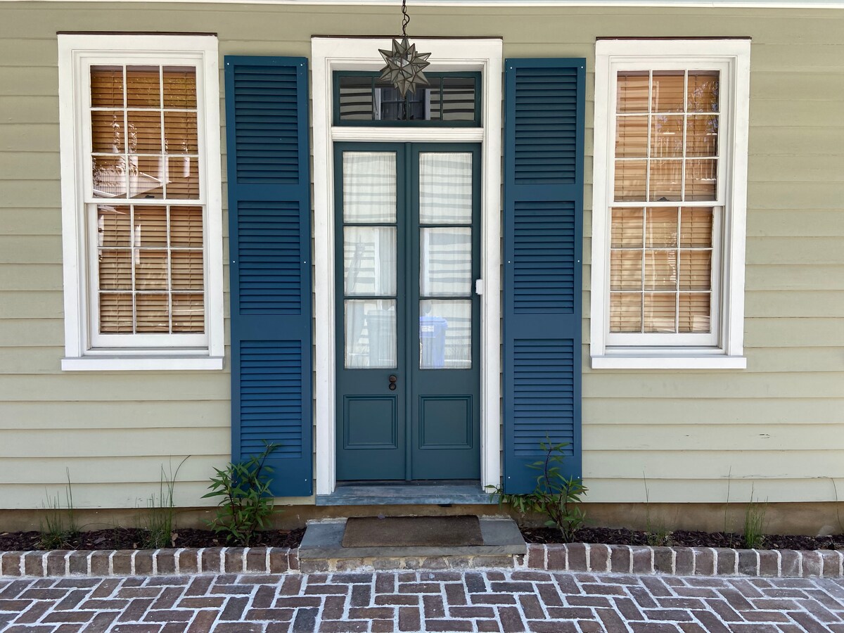 A classic entrance showcases a double door design, framed by blue shutters and set between two large windows. The brick pathway leads to the door, with small plants flanking the entrance and a decorative light fixture overhead.