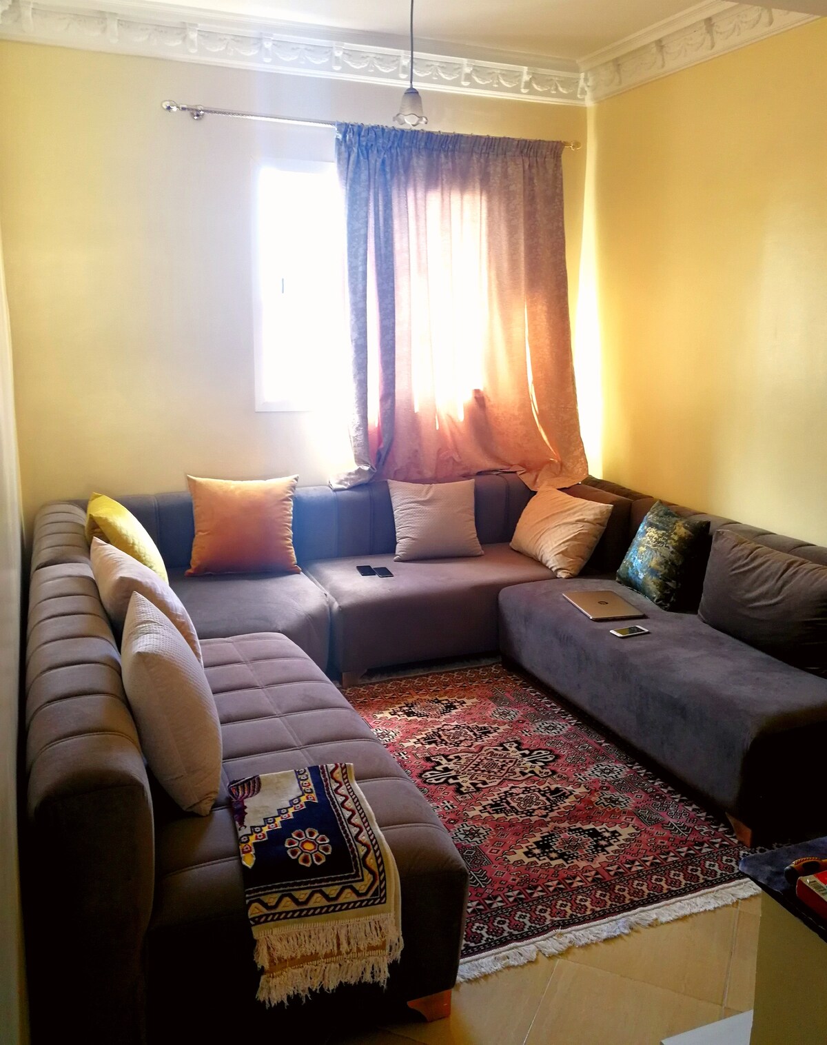 A corner living area features a spacious sectional sofa adorned with several cushions. Natural light pours in from a window draped with golden curtains, highlighting a patterned area rug beneath. Decorative elements add a touch of warmth to the inviting space.