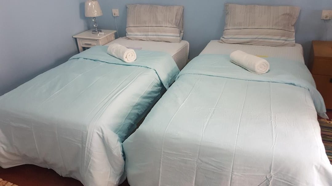The sleeping area features two super single beds, each adorned with soft, light blue coverings. Plump white towels are neatly rolled at the foot of each bed. A bedside table supports a lamp, contributing to the room's functionality.