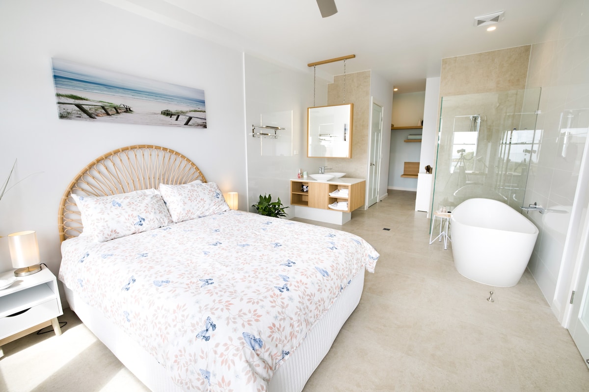 A bright bedroom features a spacious bed adorned with a floral-patterned bedspread. Natural light enters through large windows, illuminating the room. A modern ensuite is visible, showcasing a deep bathtub and a vanity area, complemented by simple decor that creates a calming environment.