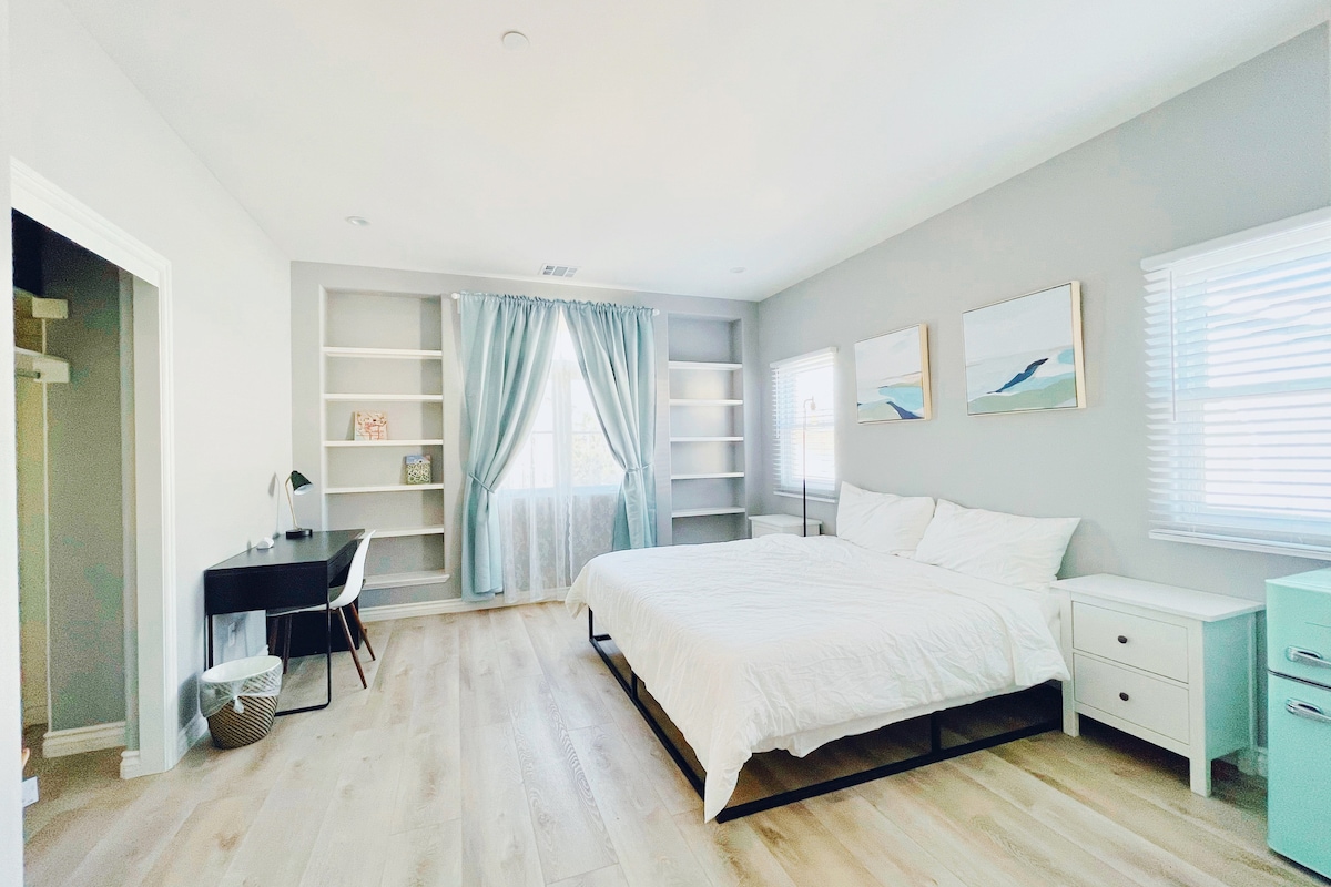 An airy bedroom features a spacious king-size memory foam bed, complemented by a dedicated workspace with a desk and chair. Light-colored walls and wood flooring create a light environment. Large windows are adorned with soft curtains, allowing natural light to brighten the room.