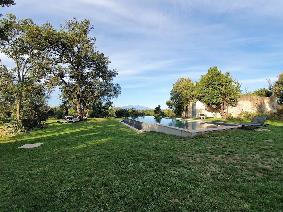 A spacious outdoor area features a shared pool surrounded by well-maintained grass and scattered trees. Lounge chairs are positioned nearby for relaxation. The picturesque landscape includes distant hills, enhancing the tranquility of the location.