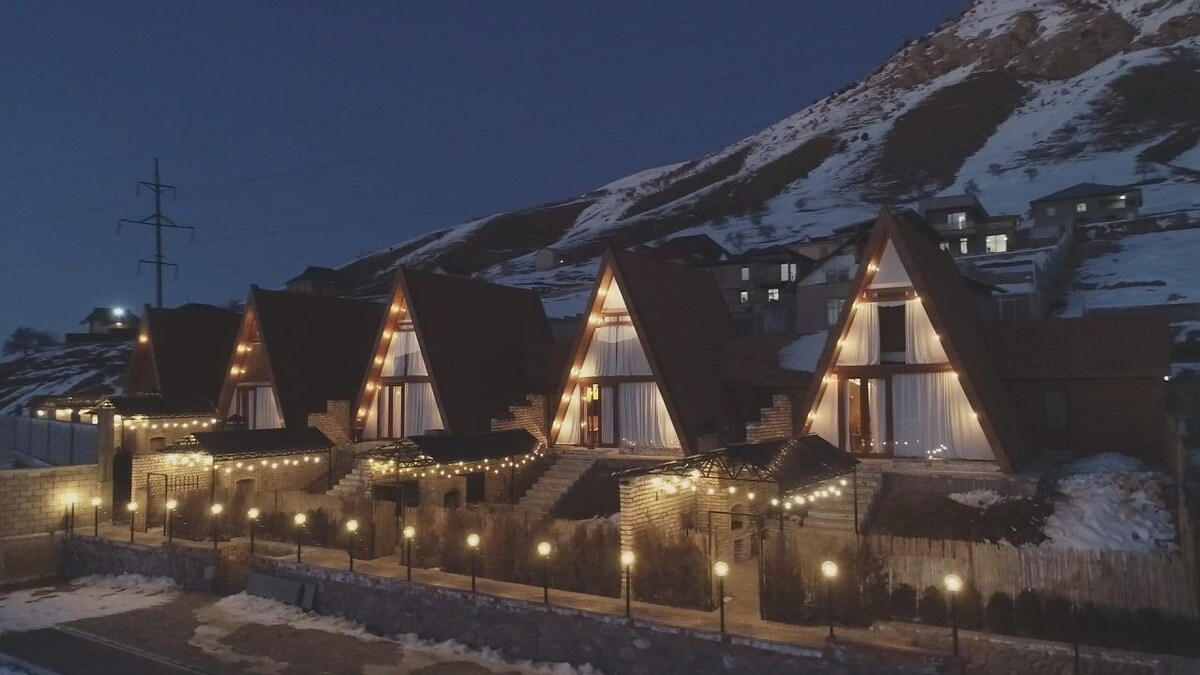 A row of A-frame chalet cottages is illuminated at night, showcasing warm lighting against a backdrop of snow-covered mountains. Each cottage features large windows framed by wooden finishes, providing a welcoming ambiance. The surrounding area is lined with softly glowing outdoor lights, enhancing the evening setting.