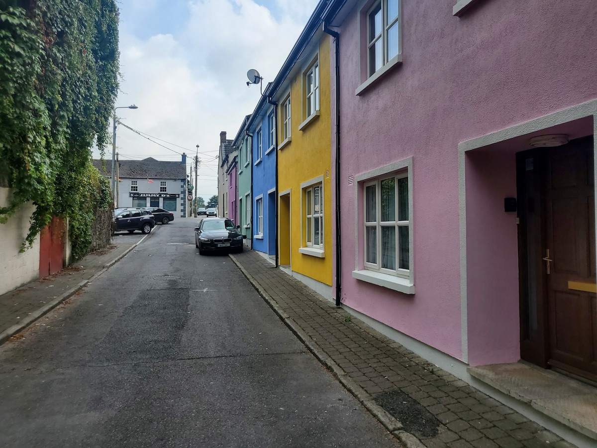 A charming street view features colorful townhouse façades in shades of yellow, pink, and blue. A parked car is positioned along the curb, with hints of greenery adorning the side of the buildings. The road is lined with cobblestones, leading into a serene residential area.