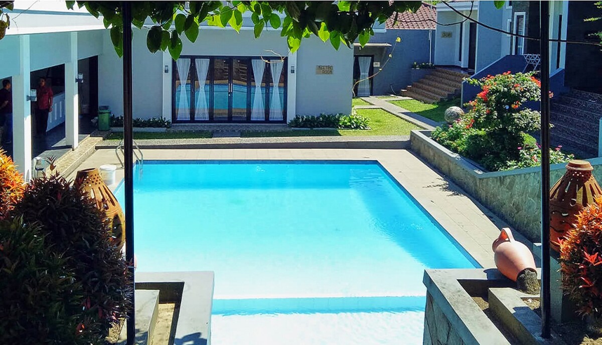 A refreshing outdoor pool is surrounded by a stone patio and lush landscaping. The pool features clear blue water and is framed by flower beds. In the background, villa buildings are visible with large windows and a well-maintained lawn extending toward the horizon.
