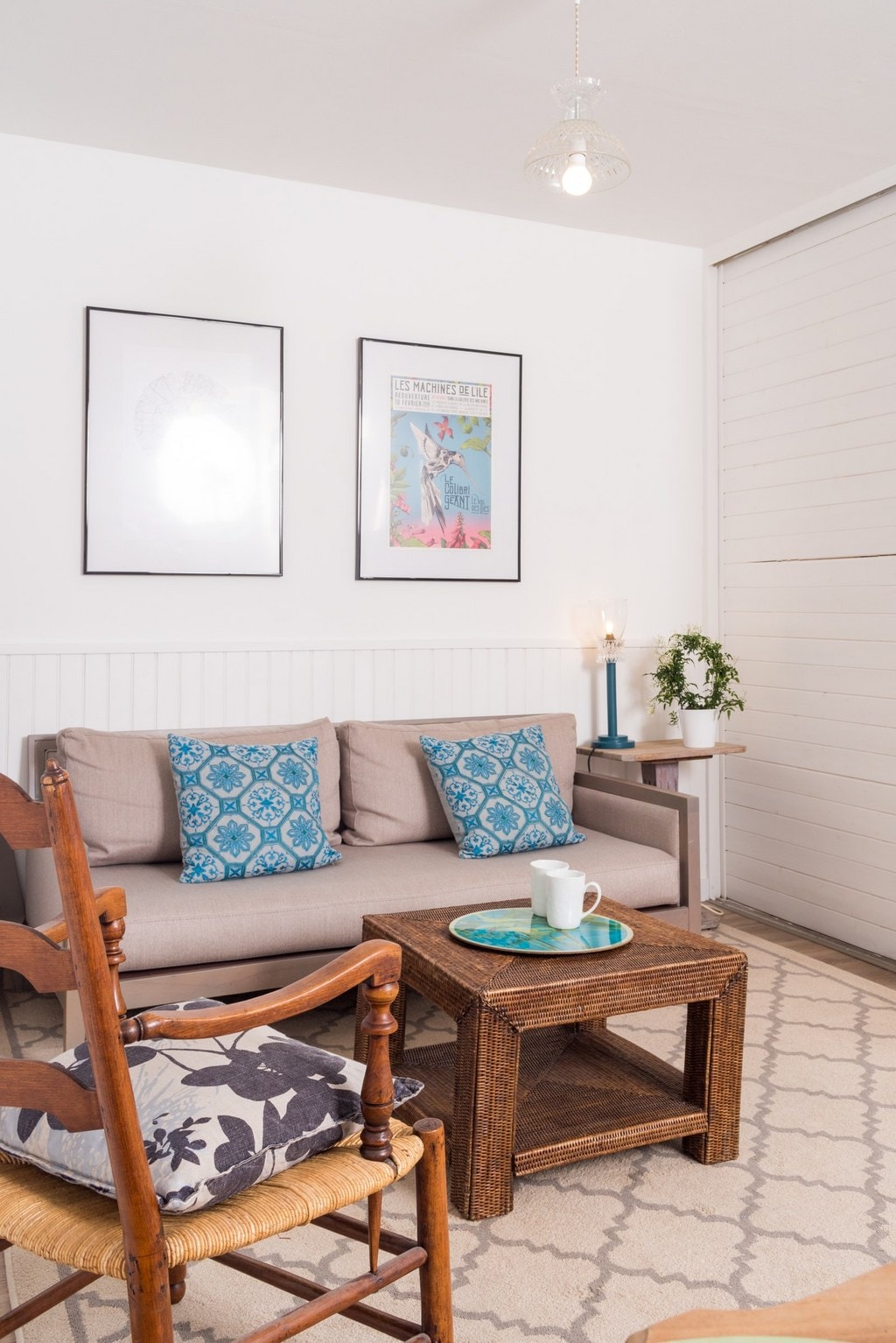 A cozy living area features a light gray sofa adorned with blue patterned cushions. A small coffee table is placed in front, accompanied by a decorative plant. Two framed artworks are hung on the wall, enhancing the inviting atmosphere of the space.