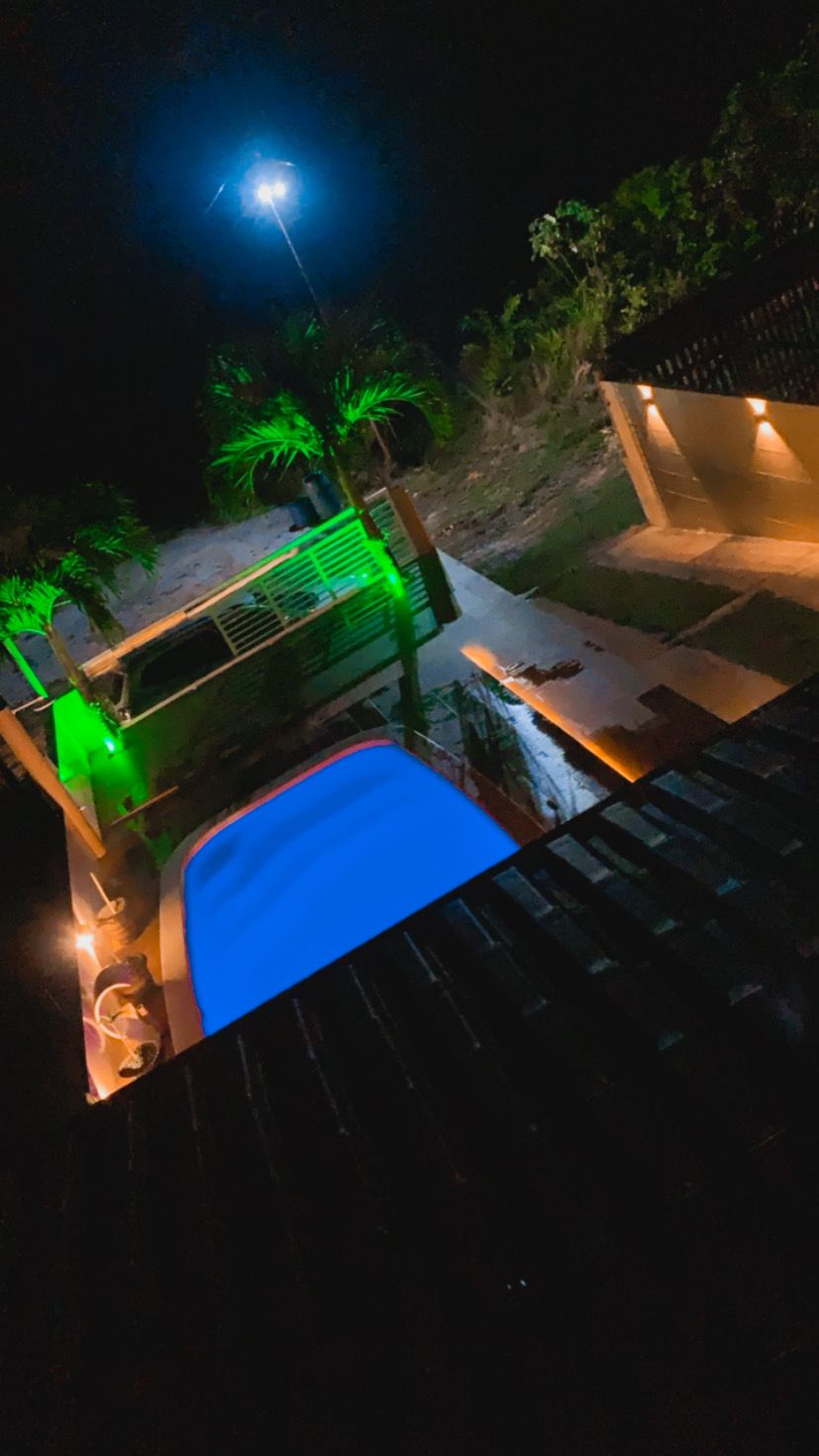 A night view of the pool area is highlighted by soft green lighting and illuminated pathways. The tranquil water reflects the surrounding greenery, while the outdoor space is enhanced by subtle ambient lighting, creating a peaceful atmosphere.