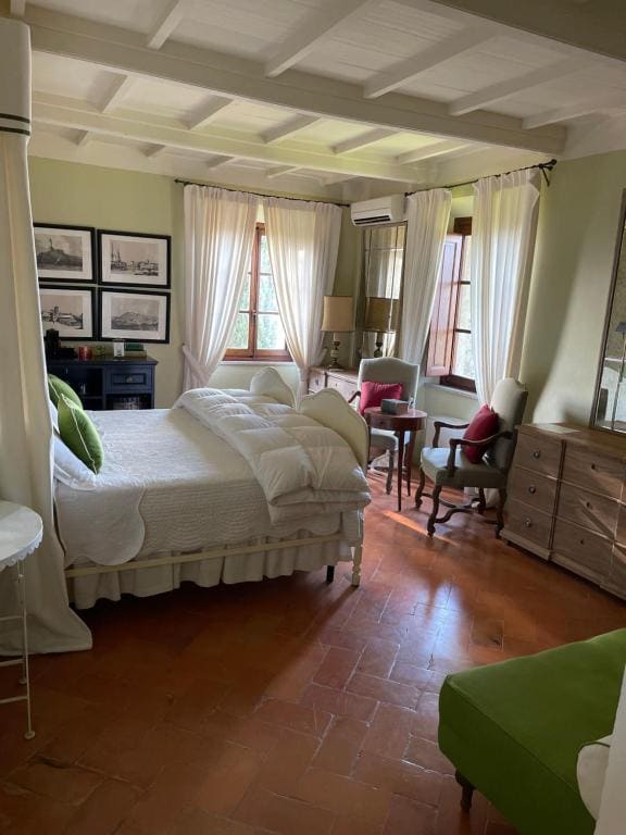 A cozy room features a spacious bed dressed in soft linens, surrounded by large windows that allow natural light to fill the space. Wooden-beam ceilings and terracotta floors add warmth. Antique furnishings, including a small desk and armchair, are present, creating a charming atmosphere.