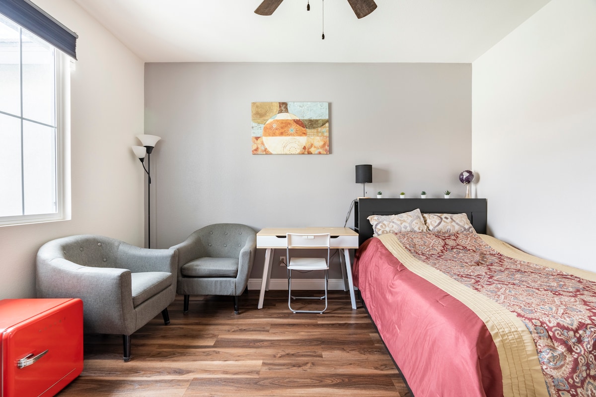A well-lit room features a spacious bed with a red and gold bedspread. A light gray armchair and a decorative table with a white chair are visible. A red storage unit adds a pop of color, while a small lamp and wall art enhance the minimalist decor.