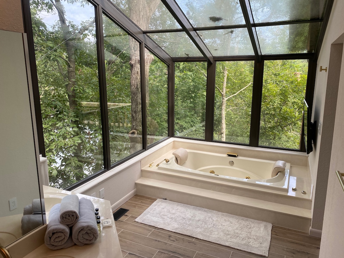 A solarium-style master bath features a jetted tub surrounded by expansive windows, offering views of the forest. Soft towels are neatly arranged on a light countertop, and natural light fills the space, enhancing the serene atmosphere.