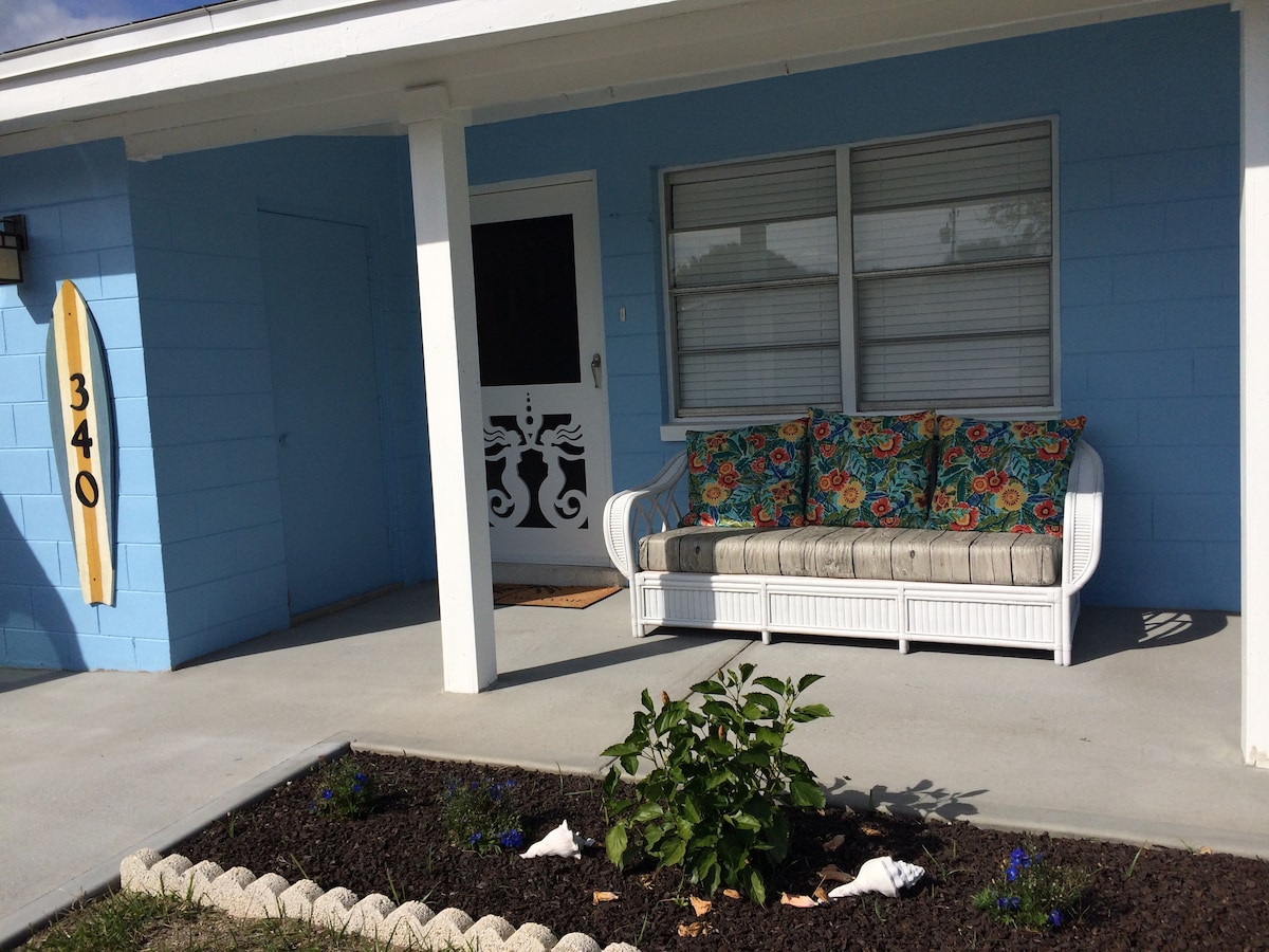 A cozy porch area is furnished with a white wicker sofa adorned with colorful cushions. A small, neatly maintained garden with flowers is visible along the walkway, while the bright blue exterior of the cottage adds a cheerful touch.