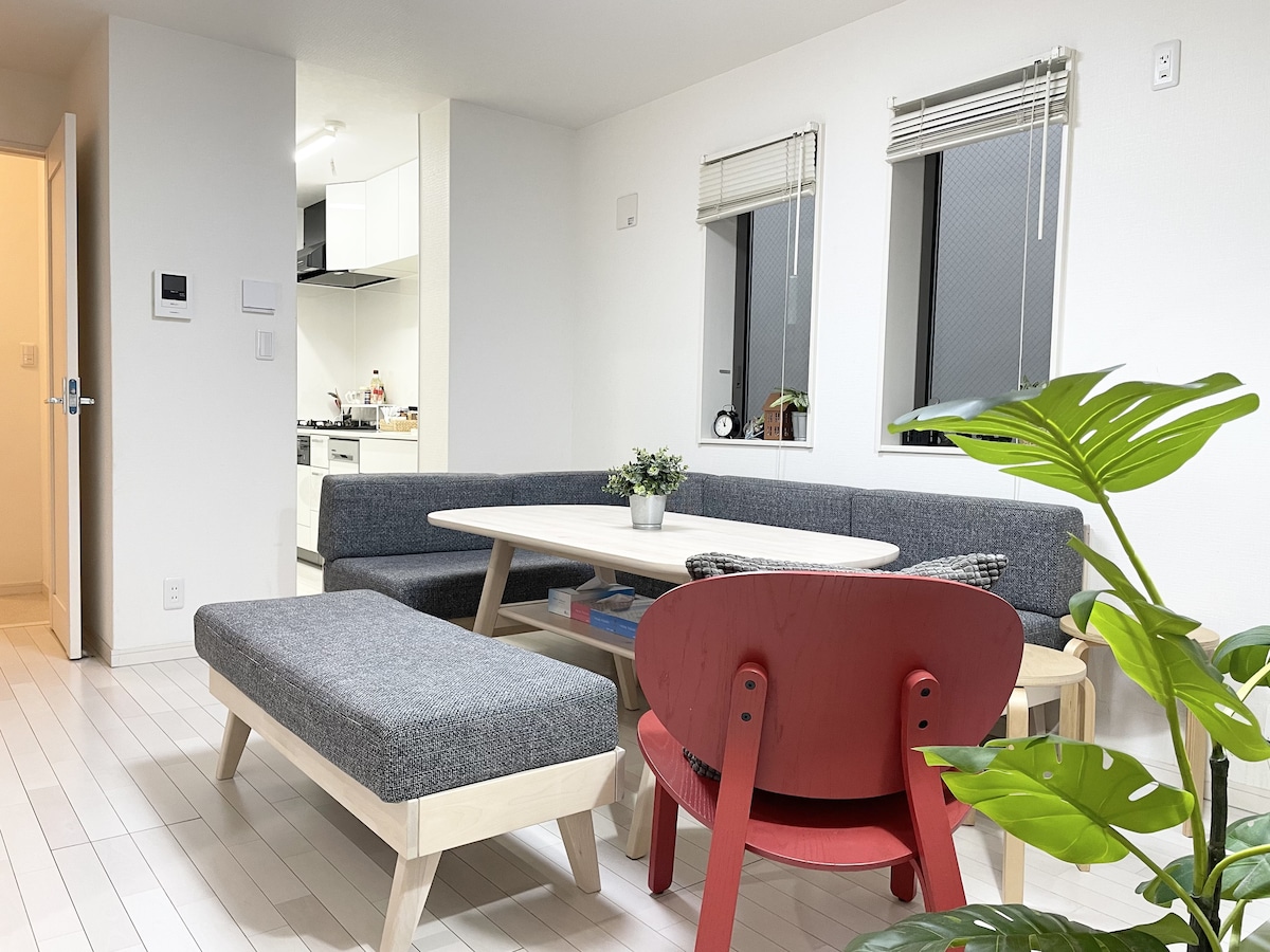 A bright living space features a gray couch and a wooden coffee table in the center. A red chair provides an accent, and plants are positioned nearby. Large windows allow natural light to fill the room, with a kitchen visible in the background.