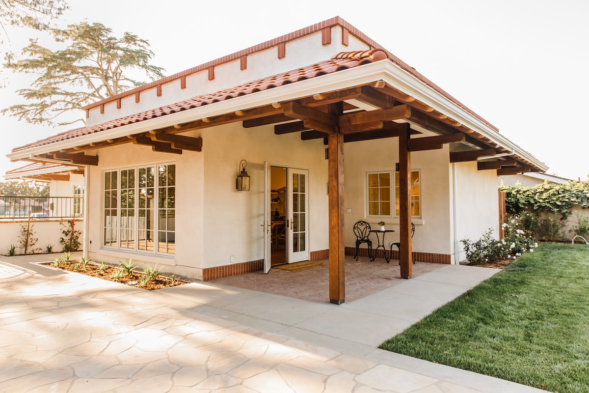 The exterior of the Casita is showcased, featuring a terracotta roof and wooden beams. A small outdoor seating area with a bistro table and two chairs invites relaxation. Lush greenery surrounds the entrance, contributing to the overall welcoming ambiance.