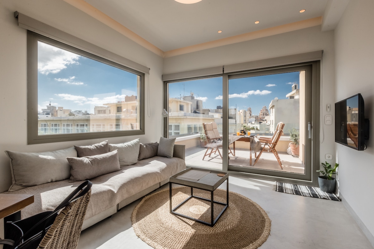A modern living area features a spacious sofa with soft cushions, complemented by a coffee table. Large glass doors reveal a bright outdoor terrace with seating, offering views of the city and clear blue sky. Natural light filters through the windows, enhancing the open feel.
