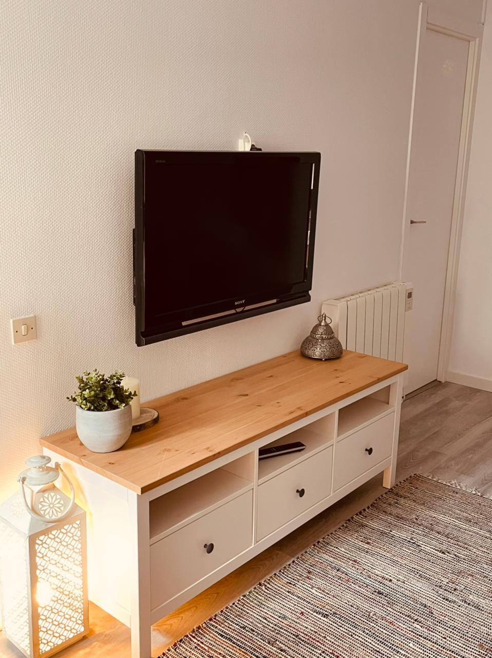 A modern living area is highlighted by a wooden media console with multiple drawers beneath a wall-mounted television. A small potted plant and a decorative lantern sit on the console. A textured rug covers the floor, complementing the light walls.