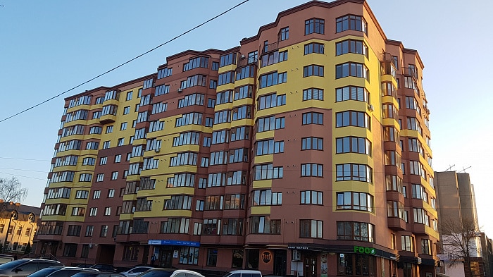 A modern multi-story building is displayed, featuring a combination of yellow and terracotta exterior. Large windows provide ample natural light to the apartments. The ground level includes retail spaces, with various signage visible. Cars are parked along the front, indicating a busy area.
