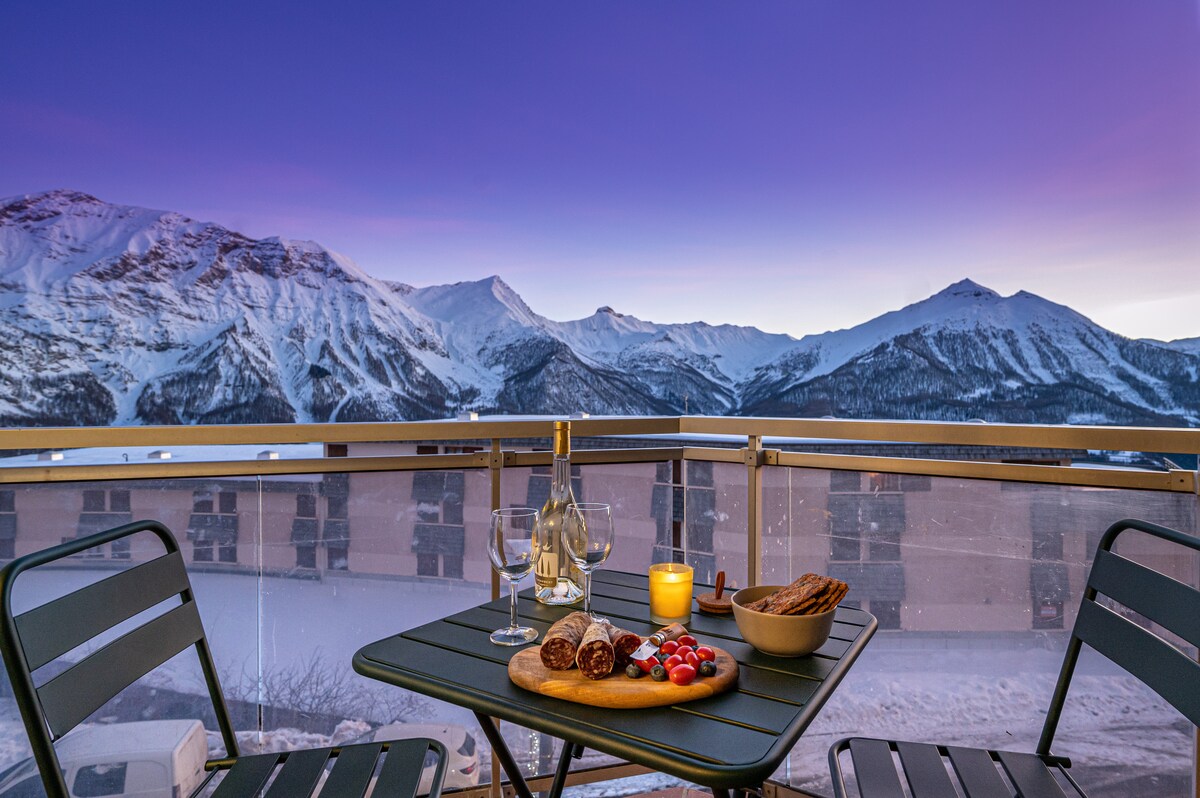 A balcony table is set with a bottle of wine, glassware, and a selection of gourmet snacks. Two folding chairs are positioned around the table. The backdrop features expansive mountain views illuminated by a purple sunset, enhancing the serene atmosphere.