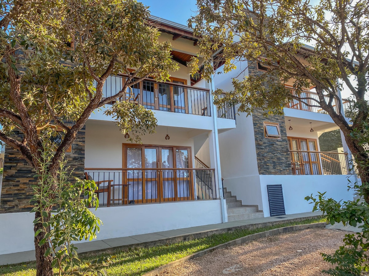 The exterior of the building is displayed, featuring two levels with balconies and large windows. Natural stone accents and wooden railings add character to the structure. A pathway lined with greenery leads up to the entrance, creating a welcoming appearance.