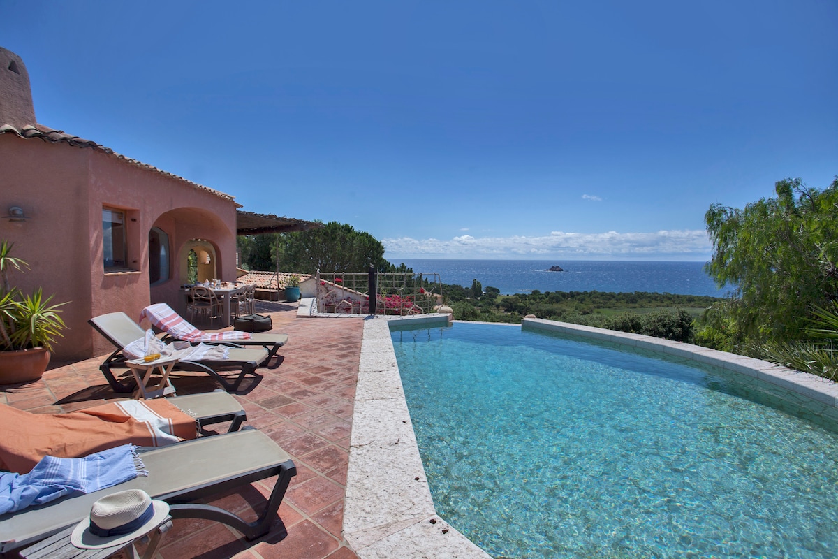 An infinity pool overlooks a serene view of the sea and distant islands. Sun loungers are arranged nearby, with a terracotta patio extending towards a shaded dining area. Lush greenery frames the property's edge, enhancing the peaceful outdoor space.