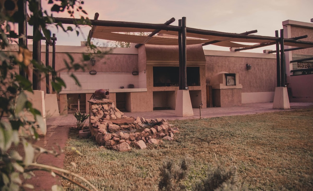 An outdoor area features a stone barbecue surrounded by grass. Wooden beams provide shade over a spacious patio area. The setting is enhanced by a warm, natural ambiance created by the evening light filtering through the trees in the background.