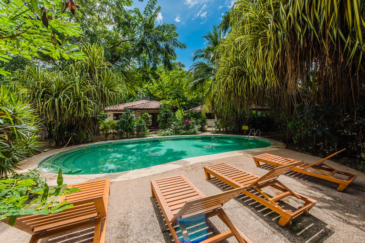 A common pool area is surrounded by lush greenery, featuring a circular pool with clear water. Several wooden lounge chairs are arranged around the pool, providing a space for relaxation, while tropical plants offer a natural ambiance.