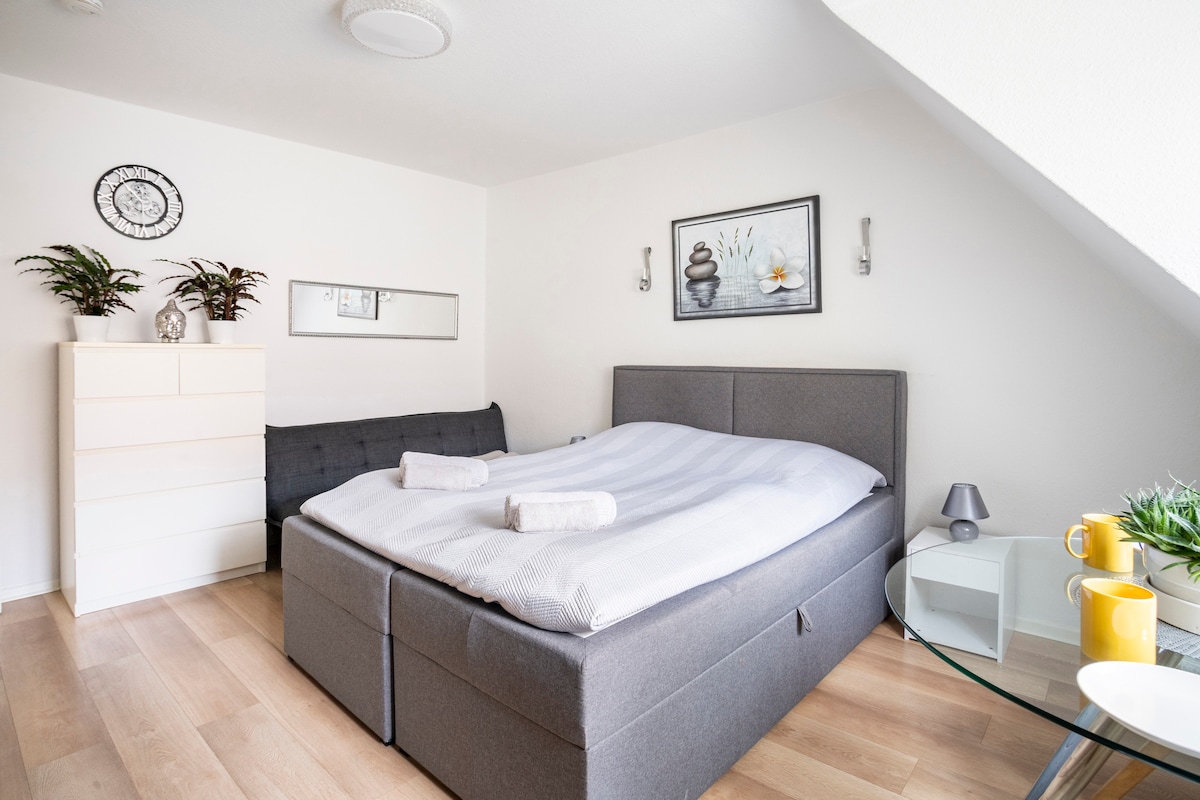 A spacious sleeping area features a gray upholstered bed with fresh linens, adorned with two rolled towels. A chest of drawers stands nearby, complemented by a mirror. Potted plants add a touch of greenery, while a small table holds a yellow cup and a decorative lamp.