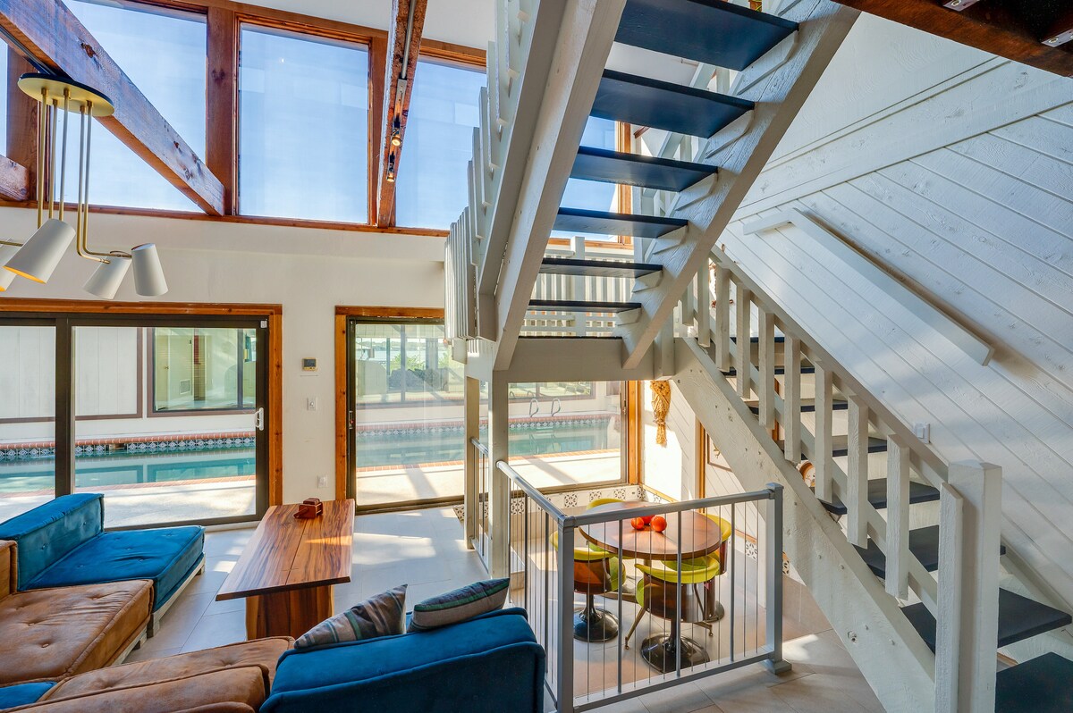 A spacious living area features large windows allowing natural light to fill the space, highlighting modern furnishings. Stairs lead to the upper level, while a wooden dining table sits next to a circular seating area. A pool can be seen through the glass doors.
