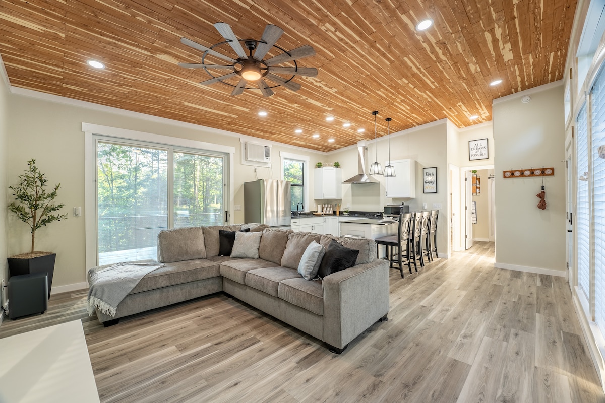 An open-concept living area features a spacious sectional couch, highlighted by natural wood ceilings and ample lighting. The kitchen showcases modern appliances, while large windows frame views of surrounding greenery. A dining area with seating for several is visible, enhancing the communal space.