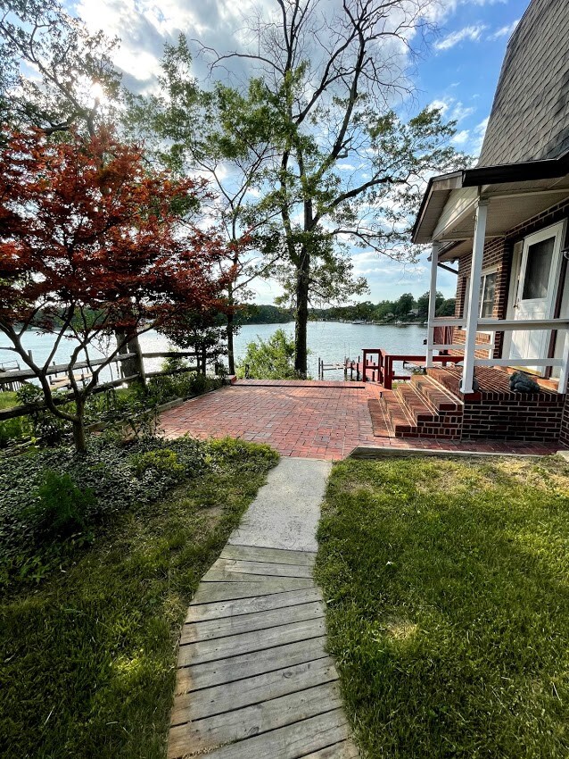 A scenic outdoor area features a brick patio leading to the water, framed by trees and landscaped greenery. The path is lined with wooden planks, offering a clear route to the waterfront. Comfortable seating is positioned to enjoy the views.