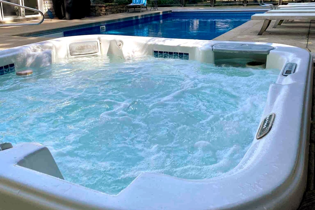 A spacious hot tub is filled with bubbling water, located adjacent to a sparkling blue pool. Surrounding the hot tub, the patio area features lounge chairs and is set in a lush, landscaped environment.