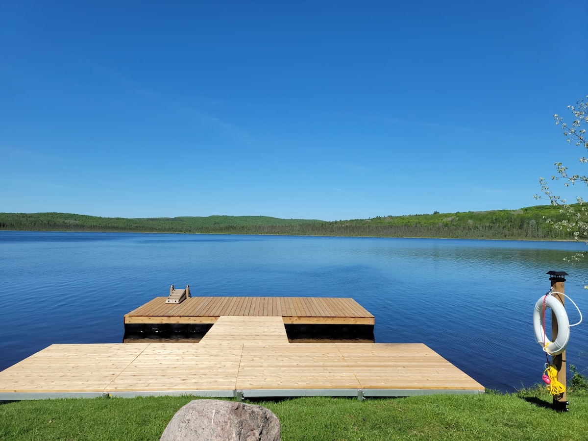 A wooden dock extends into a calm lake, surrounded by lush greenery. The clear blue sky reflects on the water's surface, creating a peaceful environment. A life ring is positioned nearby for safety, and a large rock is visible on the grassy shoreline.