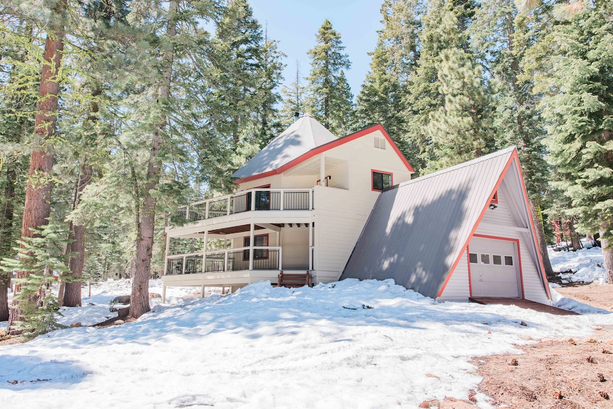 A two-story cabin is surrounded by snow-covered ground and towering pine trees. The exterior features a combination of white siding and a gray roof, with red accents highlighting the structure. A covered deck extends to the front, providing a welcoming entrance.