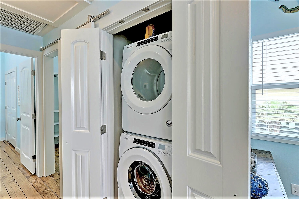 A modern laundry area is showcased, featuring a stacked washer and dryer within sleek white cabinetry. The cabinet doors are partially open, revealing the appliances. Natural light filters through a nearby window, enhancing the space's functionality.