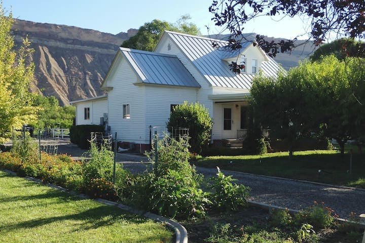 The exterior of a charming two-story home is surrounded by well-maintained gardens and greenery. A gravel pathway leads to the entrance, which is flanked by landscaping. In the background, a majestic mountain range adds to the rural scenic appeal.