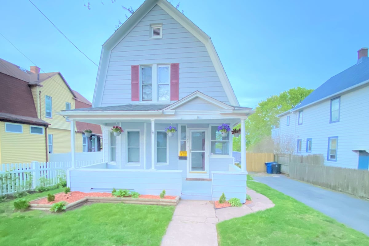 A charming Dutch colonial house features a light blue exterior with red accents. Flower boxes adorn the front windows, which are framed by a welcoming porch. A well-maintained garden with a curved pathway leads to the entrance, and neighboring houses provide a sense of community.