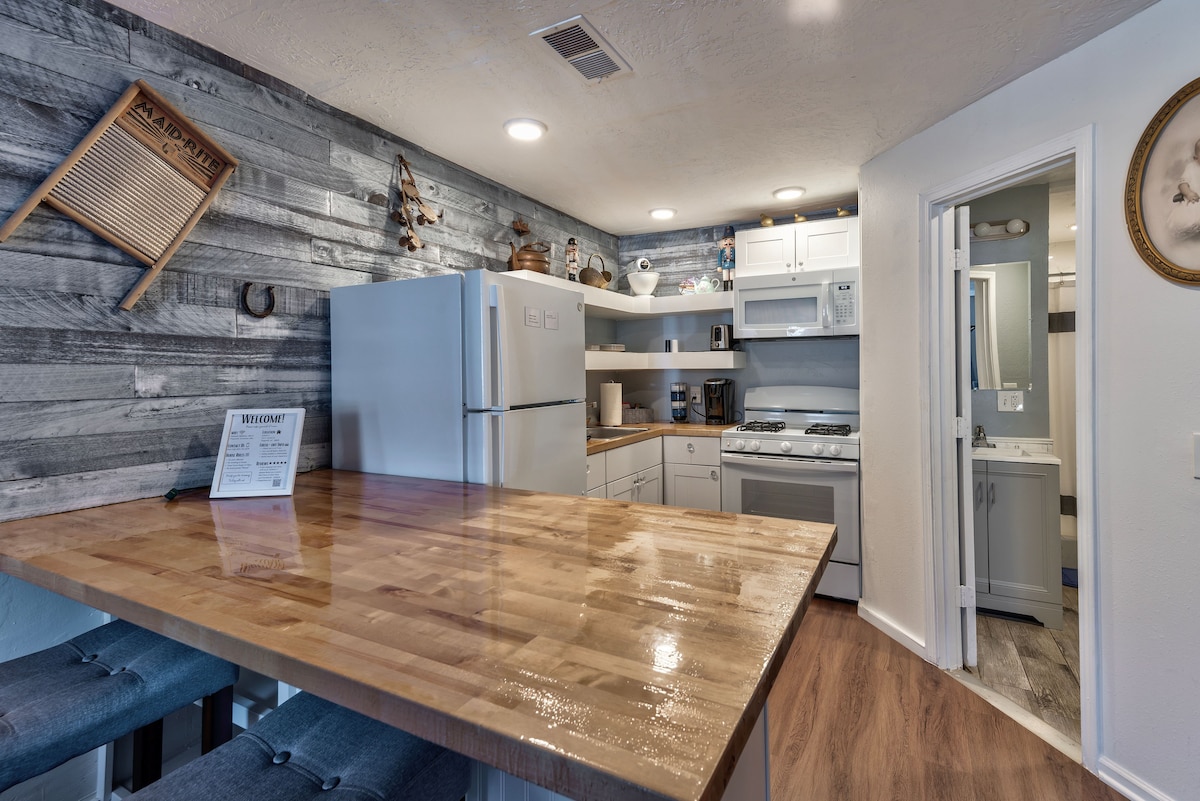 A modern kitchen is presented with wooden cabinetry and a polished countertop. Appliances include a refrigerator and a gas stove. A small dining area features stools set at the countertop. Light fixtures brighten the space, while decorative items adorn the walls.