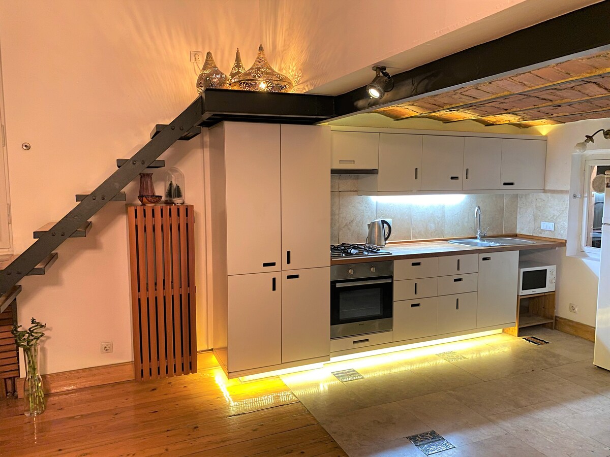 A modern kitchen is displayed with streamlined cabinetry, complemented by warm lighting that highlights the countertops. Essential appliances, such as a stove, kettle, and microwave, are visible. The kitchen is situated beneath a wooden beam ceiling, with a staircase leading to an upper level.