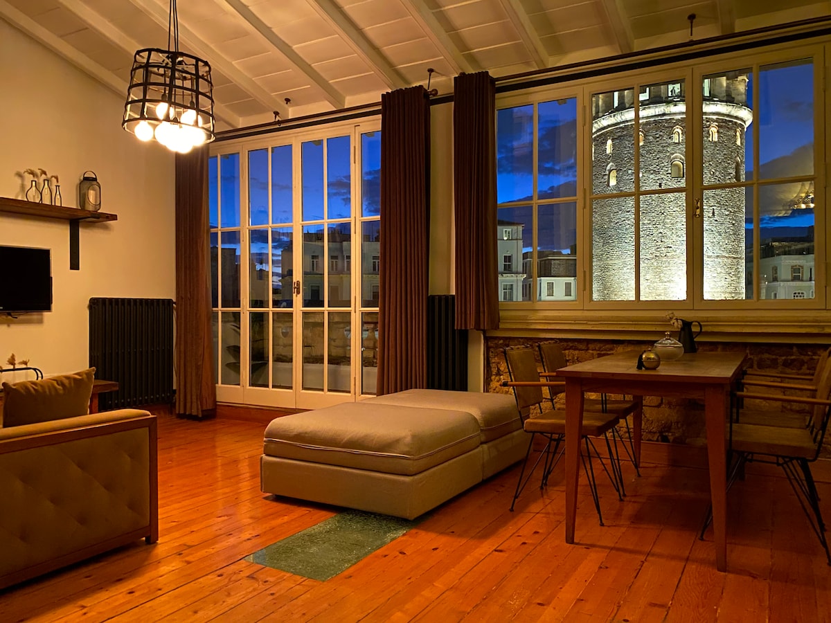 A spacious living area features a large window showcasing views of the illuminated Galata Tower against a twilight sky. Wooden floors complement the room, which includes a cozy sofa, a coffee table, and a dining table with chairs, providing a welcoming space for relaxation.