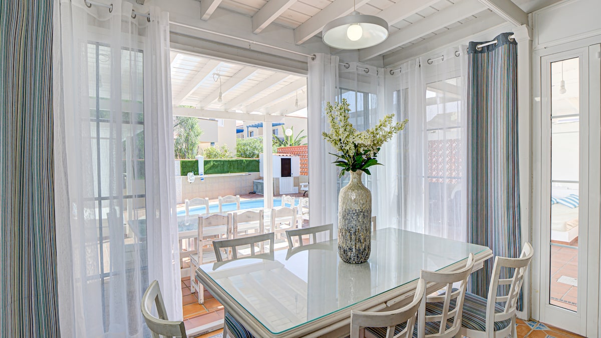 A bright dining area features a glass table surrounded by light-colored chairs. Sheer curtains frame the windows, allowing natural light to illuminate the space. A floral centerpiece adds a touch of life, with views of a private pool visible through the open doors.