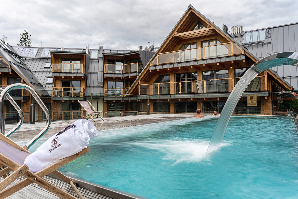 An outdoor heated pool features a gentle water flow and is surrounded by wooden deck chairs. The building showcases a distinctive architectural design with multiple balconies and large glass panels, allowing for expansive views of the area.