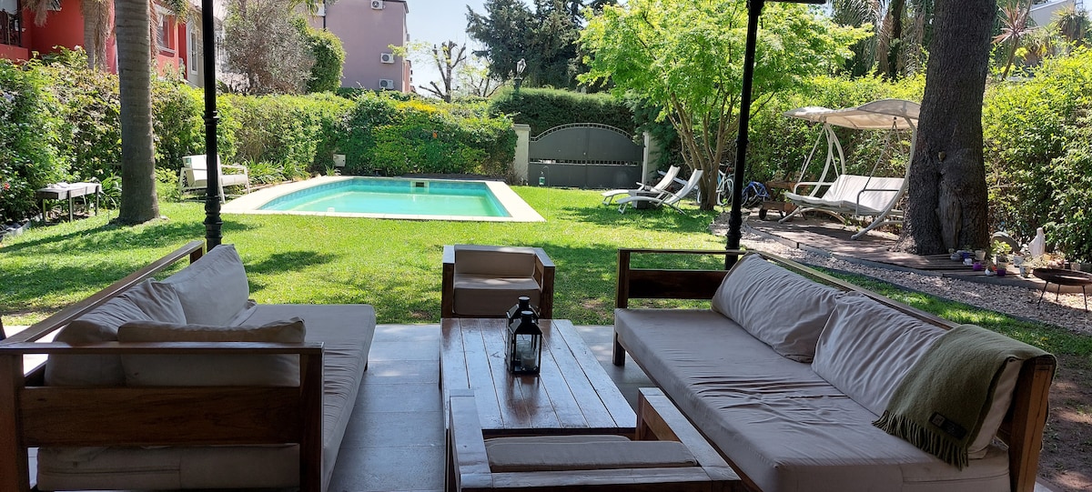 A shaded outdoor seating area is presented with wooden sofas and a central table. Lush green grass surrounds a rectangular pool in the background, accompanied by sun loungers and a relaxing swing. Mature trees provide additional shade and a sense of privacy.