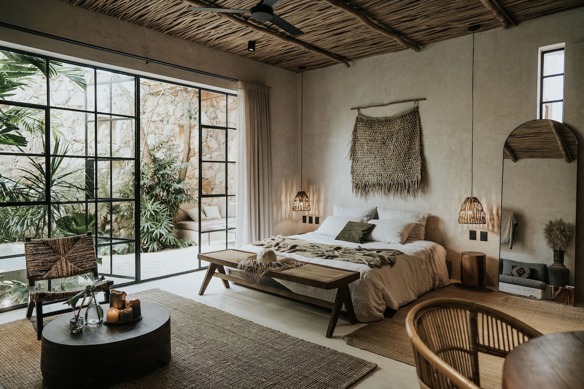 A calming bedroom is featured with natural textures and earthy tones. A spacious bed is complemented by woven wall art and pendant lights. Large glass doors provide a view of tropical greenery, while seating and decor maintain a harmonious aesthetic.