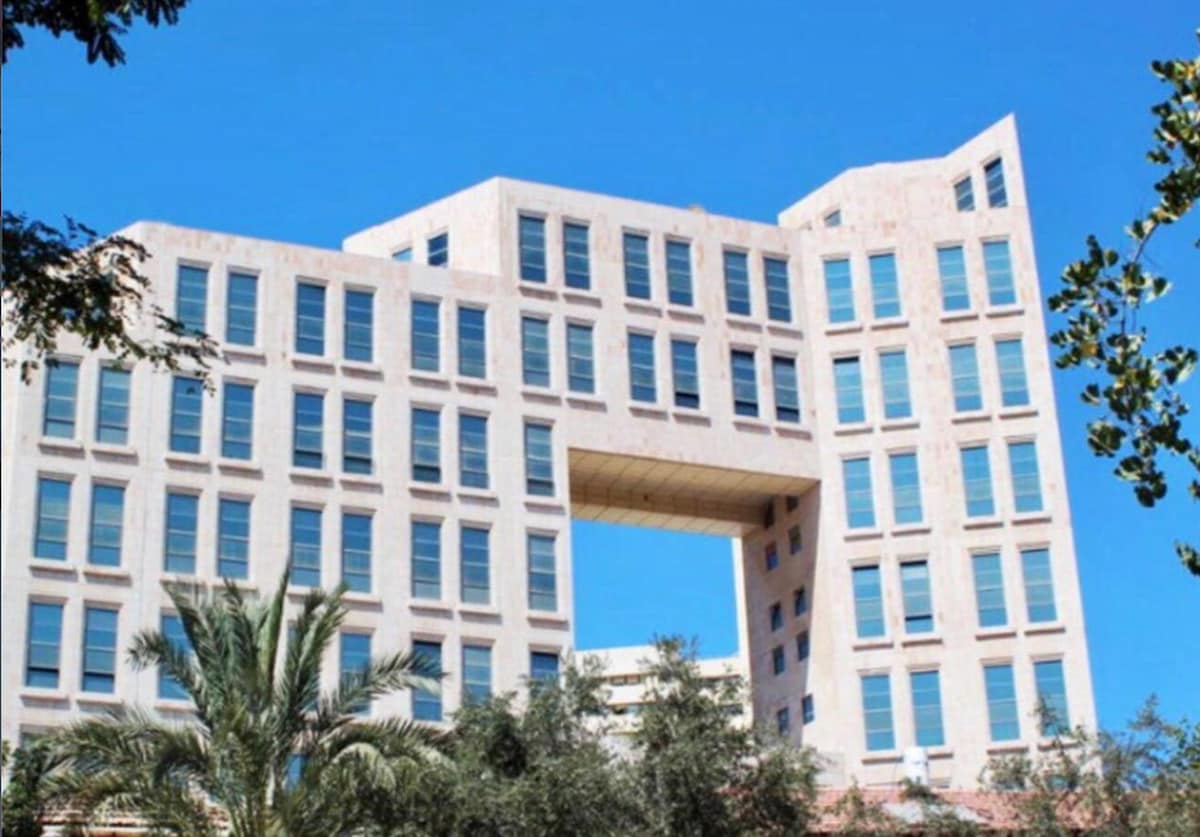 A modern building with a unique architectural design is depicted, featuring a central opening framed by large glass windows. Lush palm trees are visible in the foreground, complementing the bright blue sky that serves as the backdrop.
