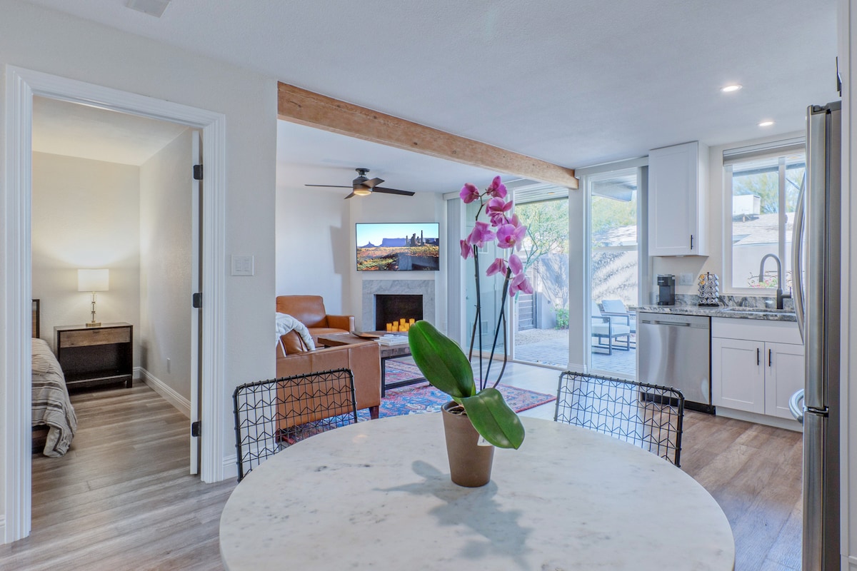 A light-filled room is shown, featuring a round dining table at the forefront with a potted plant. In the background, an open area includes a cozy seating arrangement and kitchen with modern appliances. Double doors lead to a serene outdoor space.