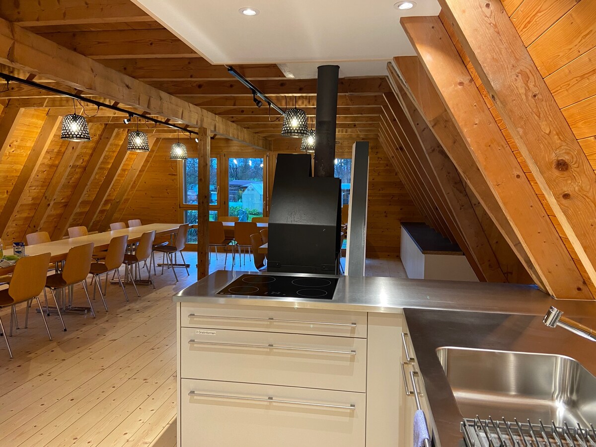A modern kitchen area is showcased, featuring a sleek stove and stainless steel sink. The wooden ceiling beams complement the open dining space in the background, where a large table is arranged with multiple chairs, inviting group gatherings and meals.